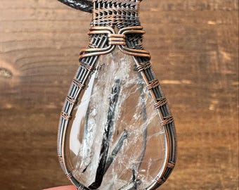 May include: A teardrop-shaped pendant featuring a clear quartz crystal with black inclusions, encased in a copper-colored wire wrap. The pendant hangs from a black cord, showcasing a handcrafted jewelry design.