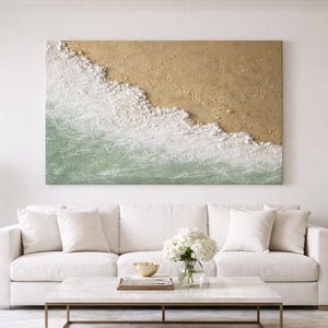 Könnte beinhalten: Ein großes, rechteckiges abstraktes Gemälde, das eine Strandszene mit strukturiertem Sand, schäumenden weißen Wellen und grünem Wasser darstellt. Das Kunstwerk hängt über einem weißen Sofa mit weißen Kissen. Zwei weiße Lampen stehen auf Beistelltischen.