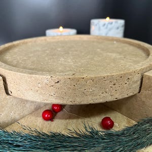 May include: A beige stone serving tray with a round top and a pedestal base. The tray is decorated with red berries and teal-coloured decorative grass. Two lit candles are in the background.