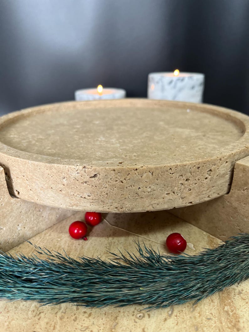 May include: A beige stone serving tray with a round top and a pedestal base. The tray is decorated with red berries and teal-coloured decorative grass. Two lit candles are in the background.