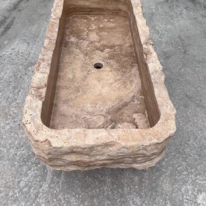 May include: A rectangular stone sink featuring a natural, rough-hewn exterior with layered, textured beige and tan surfaces. The interior basin is smooth with a polished finish and includes a single circular drain hole centred at the bottom.