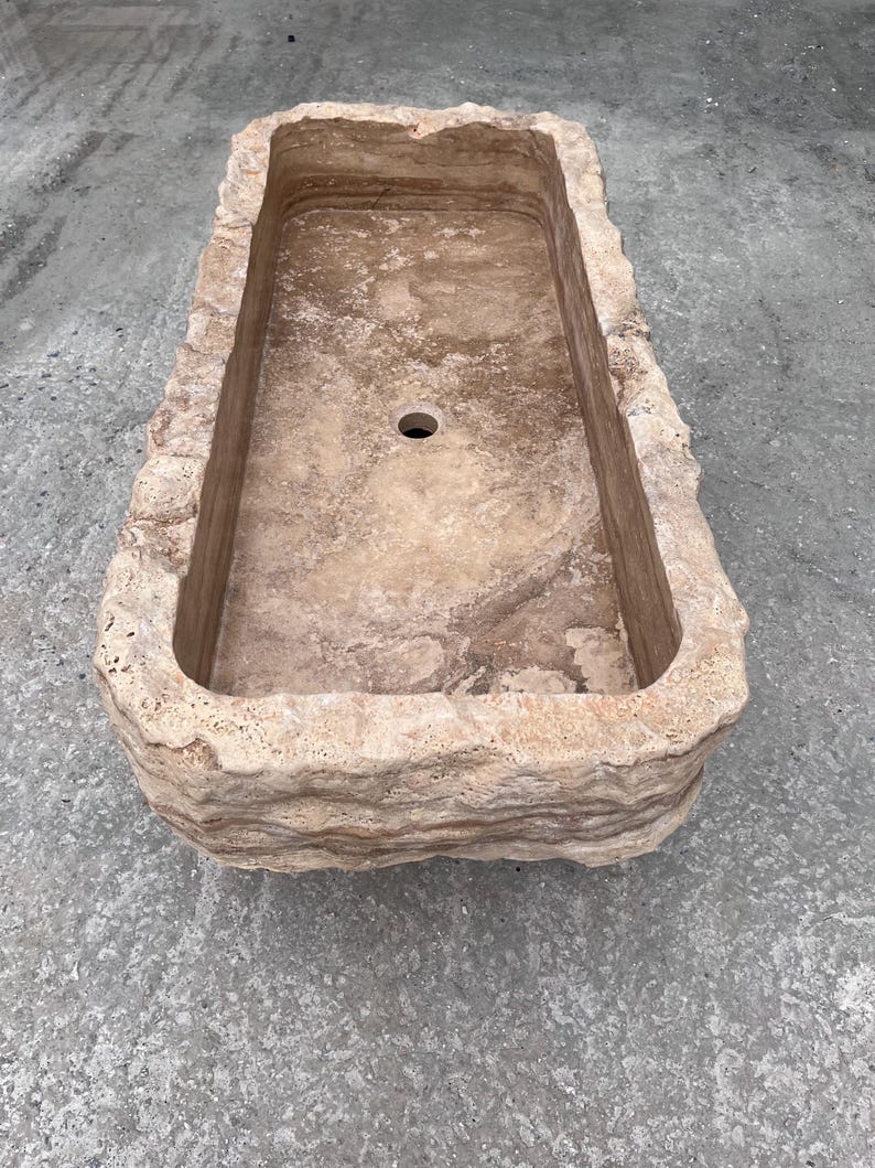 May include: A rectangular stone sink featuring a natural, rough-hewn exterior with layered, textured beige and tan surfaces. The interior basin is smooth with a polished finish and includes a single circular drain hole centred at the bottom.