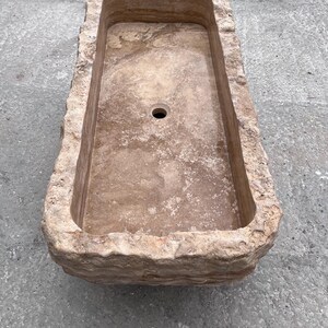 May include: A rectangular stone sink with a natural, rough-hewn exterior and a smooth, polished interior basin. The sink features a light tan and beige colour palette with visible sedimentary layers and a single circular drain hole in the centre.