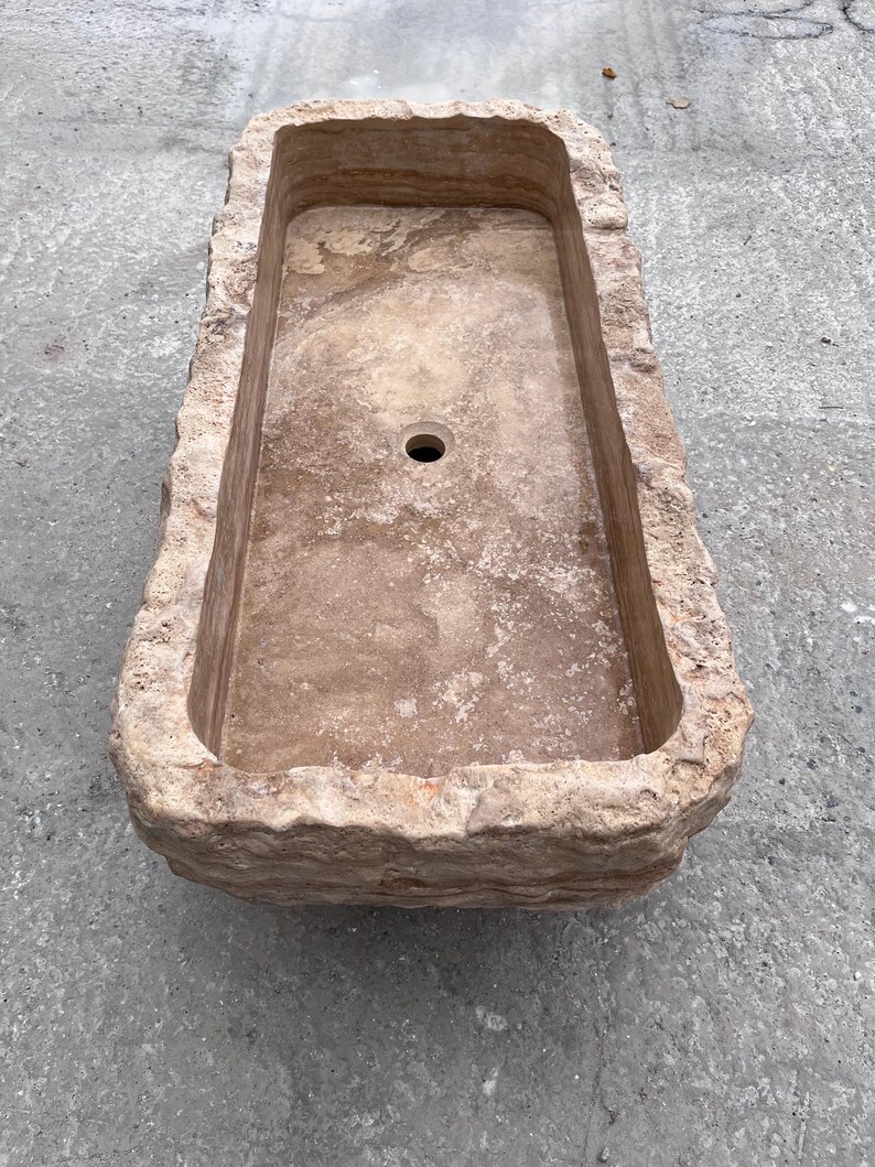 May include: A rectangular stone sink with a natural, rough-hewn exterior and a smooth, polished interior basin. The sink features a light tan and beige colour palette with visible sedimentary layers and a single circular drain hole in the centre.