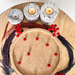 May include: A decorative arrangement featuring three marble candle holders with lit candles, a round tray, and red berries. The candle holders are white with grey veining. The tray is beige, and the berries are bright red. Colourful dried plants surround the tray.