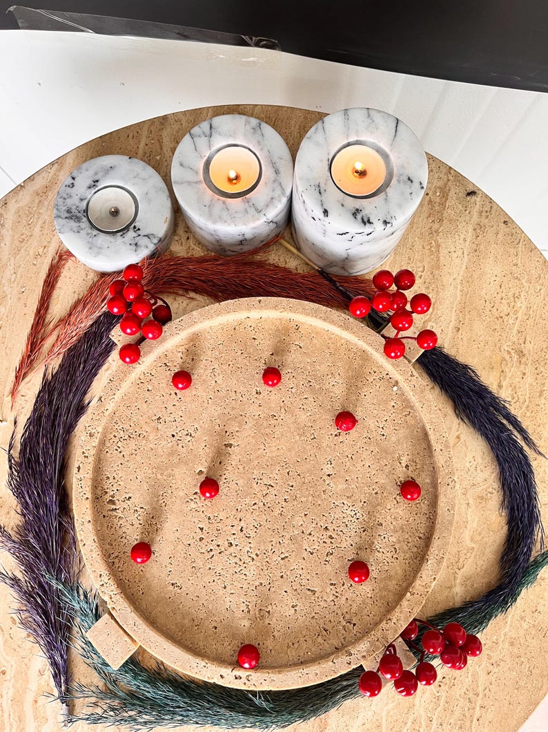 May include: A decorative arrangement featuring three marble candle holders with lit candles, a round tray, and red berries. The candle holders are white with grey veining. The tray is beige, and the berries are bright red. Colourful dried plants surround the tray.