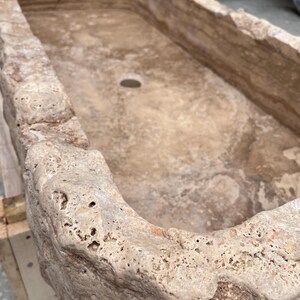 May include: A rectangular stone sink carved from beige travertine with a rough, porous, and natural rock-like exterior texture. The interior basin is smooth with visible sedimentary layers and features a single circular drain hole at the centre.