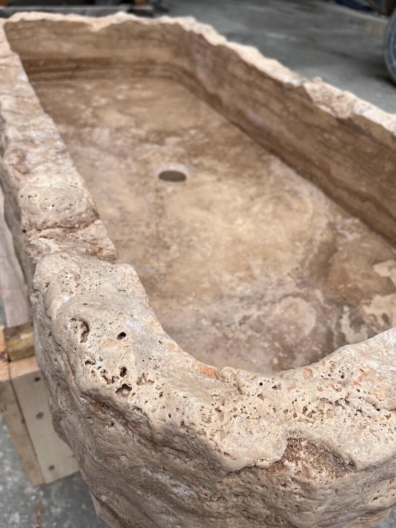 May include: A rectangular stone sink carved from beige travertine with a rough, porous, and natural rock-like exterior texture. The interior basin is smooth with visible sedimentary layers and features a single circular drain hole at the centre.