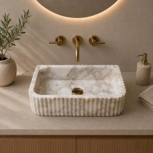 Op de afbeelding: Een rechthoekige wastafel van wit marmer met een geribbelde buitenkant en een gepolijste afwerking. De waskom heeft grijze aders en een goudkleurige afvoer. Het is gemonteerd op een aanrechtblad met goudkleurige wandkranen.