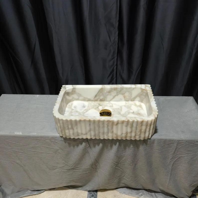May include: A rectangular stone sink featuring a white and grey marbled pattern with a fluted exterior texture. The basin has a smooth interior surface and includes a centered circular gold-toned drain fixture at the bottom of the vessel.