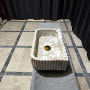 May include: A rectangular marble sink featuring a white and beige mottled stone pattern with a fluted exterior texture. The basin includes a centered, circular, polished gold-toned drain cover, providing a distinct contrast to the natural stone finish.