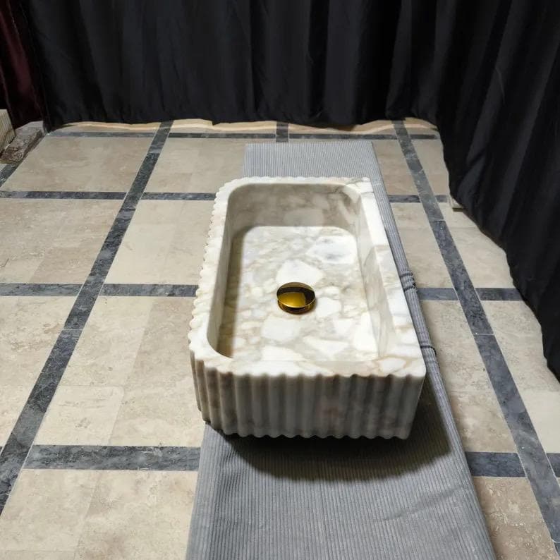 May include: A rectangular marble sink featuring a white and beige mottled stone pattern with a fluted exterior texture. The basin includes a centered, circular, polished gold-toned drain cover, providing a distinct contrast to the natural stone finish.