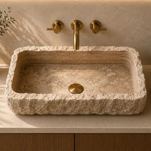 May include: A rectangular bathroom sink carved from beige natural stone with a rough, chiseled exterior texture and a smooth interior basin. It features a centred circular drain cover in a brushed brass finish, matching the wall-mounted faucet.