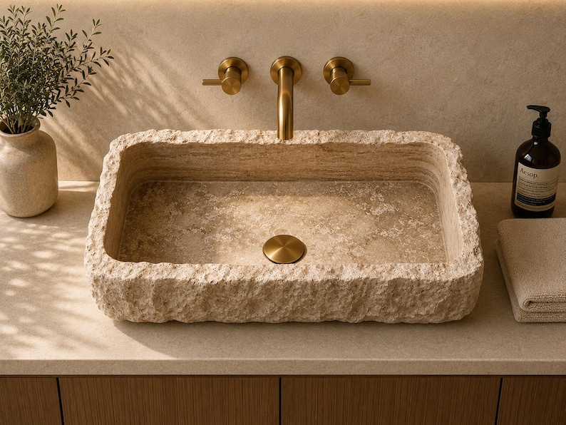 May include: A rectangular bathroom sink carved from beige natural stone with a rough, chiseled exterior texture and a smooth interior basin. It features a centred circular drain cover in a brushed brass finish, matching the wall-mounted faucet.