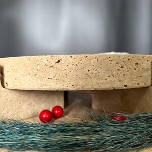 May include: A light brown, porous, rectangular object with a cutout, possibly a display stand. Two small, shiny red spheres and a sprig of blue-green foliage are in front of the object, suggesting a festive theme.