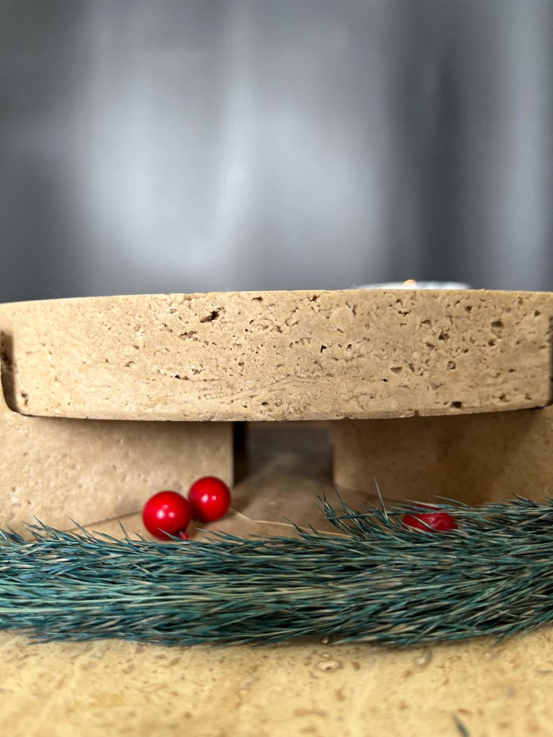 May include: A light brown, porous, rectangular object with a cutout, possibly a display stand. Two small, shiny red spheres and a sprig of blue-green foliage are in front of the object, suggesting a festive theme.
