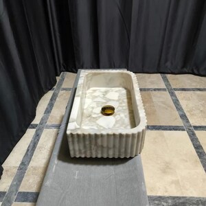 May include: A rectangular stone sink featuring a white and cream-colored terrazzo-style pattern with a fluted exterior texture. The basin includes a centered, circular, polished gold-toned drain cover, providing a contrast to the light stone surface.