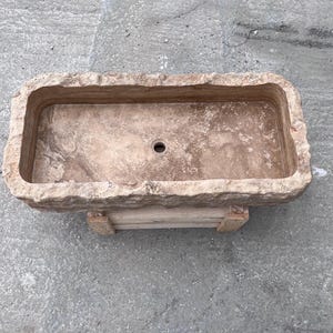 May include: A rectangular stone sink with a rough, chiseled exterior edge and a smooth, polished interior basin. The natural tan and beige stone features visible veining and a single centred circular drain hole, resting on a wooden support base.