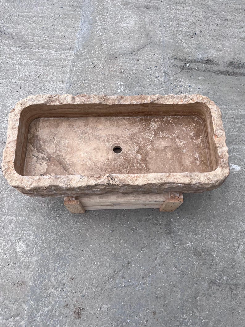 May include: A rectangular stone sink with a rough, chiseled exterior edge and a smooth, polished interior basin. The natural tan and beige stone features visible veining and a single centred circular drain hole, resting on a wooden support base.