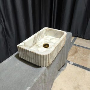 May include: A rectangular bathroom sink crafted from white marble with grey veining. The exterior features a vertical fluted texture along the sides. A circular, polished gold-tone metal drain cover is centered at the bottom of the basin.