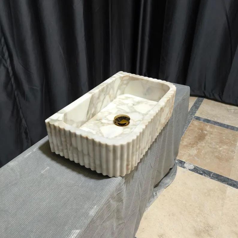 May include: A rectangular bathroom sink crafted from white marble with grey veining. The exterior features a vertical fluted texture along the sides. A circular, polished gold-tone metal drain cover is centered at the bottom of the basin.