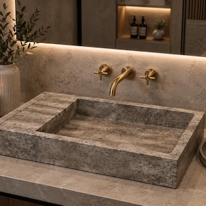 May include: A rectangular stone bathroom sink with a mottled gray and brown textured surface. The sink features a flat side ledge and is paired with a wall-mounted gold-toned faucet with two cross-handle controls, set against a matching stone wall.