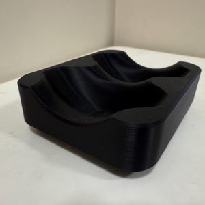 May include: A black, rectangular object with curved indentations. The item appears to be made of a solid material and has a smooth, matte finish. The design suggests it may be a holder for multiple items.