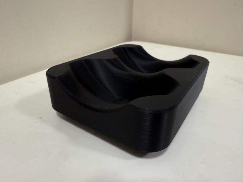 May include: A black, rectangular object with curved indentations. The item appears to be made of a solid material and has a smooth, matte finish. The design suggests it may be a holder for multiple items.