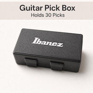 May include: A rectangular black guitar pick box with a textured lid featuring the white Ibanez logo. The box has two front latches and text above it stating Guitar Pick Box and Holds 30 Picks. The container is designed for storage.