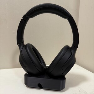 May include: Black over-ear headphones with a padded headband and earcups, resting on a black charging stand. The headphones have a sleek, modern design, perfect for listening to music or podcasts.