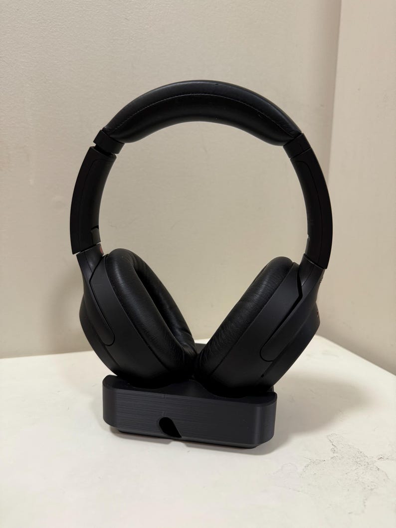 May include: Black over-ear headphones with a padded headband and earcups, resting on a black charging stand. The headphones have a sleek, modern design, perfect for listening to music or podcasts.