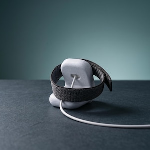 May include: A light gray speckled charging stand for a smartwatch, featuring a smooth, rounded base with a central cutout. A dark gray woven fabric watch band is wrapped around the stand, and a white charging cable extends from the center.