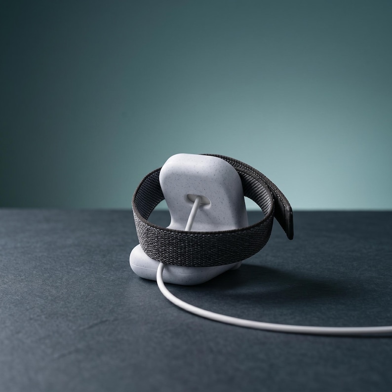 May include: A light gray speckled charging stand for a smartwatch, featuring a smooth, rounded base with a central cutout. A dark gray woven fabric watch band is wrapped around the stand, and a white charging cable extends from the center.