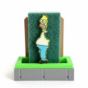 Puede incluir: Un soporte de esponja de cocina novedoso con una imagen de dibujos animados de Homer Simpson. La esponja verde para fregar se sujeta con un soporte de plástico verde y gris. La imagen de Homer Simpson es amarilla, blanca, azul y negra.