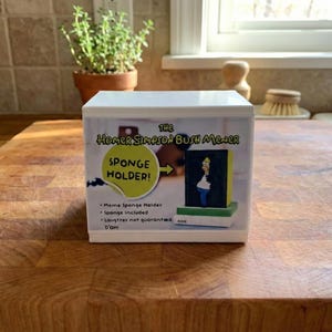 May include: A white sponge holder with the text "The Homer Simpson Bush Mower" and "Sponge Holder!" in yellow. The holder features a small image of Homer Simpson. The holder is on a wooden surface.