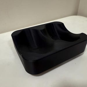 May include: A black, rectangular tray with two curved indentations. The tray is made of a solid, matte material and sits on a white surface. The design suggests it could be used to hold small items.