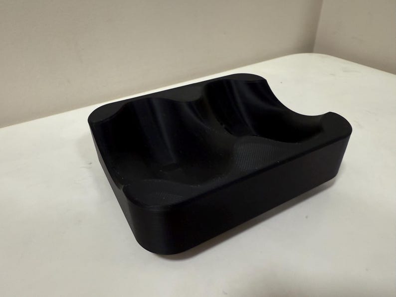 May include: A black, rectangular tray with two curved indentations. The tray is made of a solid, matte material and sits on a white surface. The design suggests it could be used to hold small items.