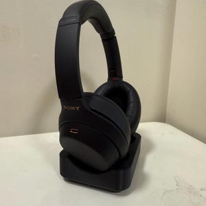 May include: Black over-ear headphones with the brand name "SONY" in gold lettering. The headphones are on a black charging stand. The earcups are round and padded. The headphones are on a white surface.