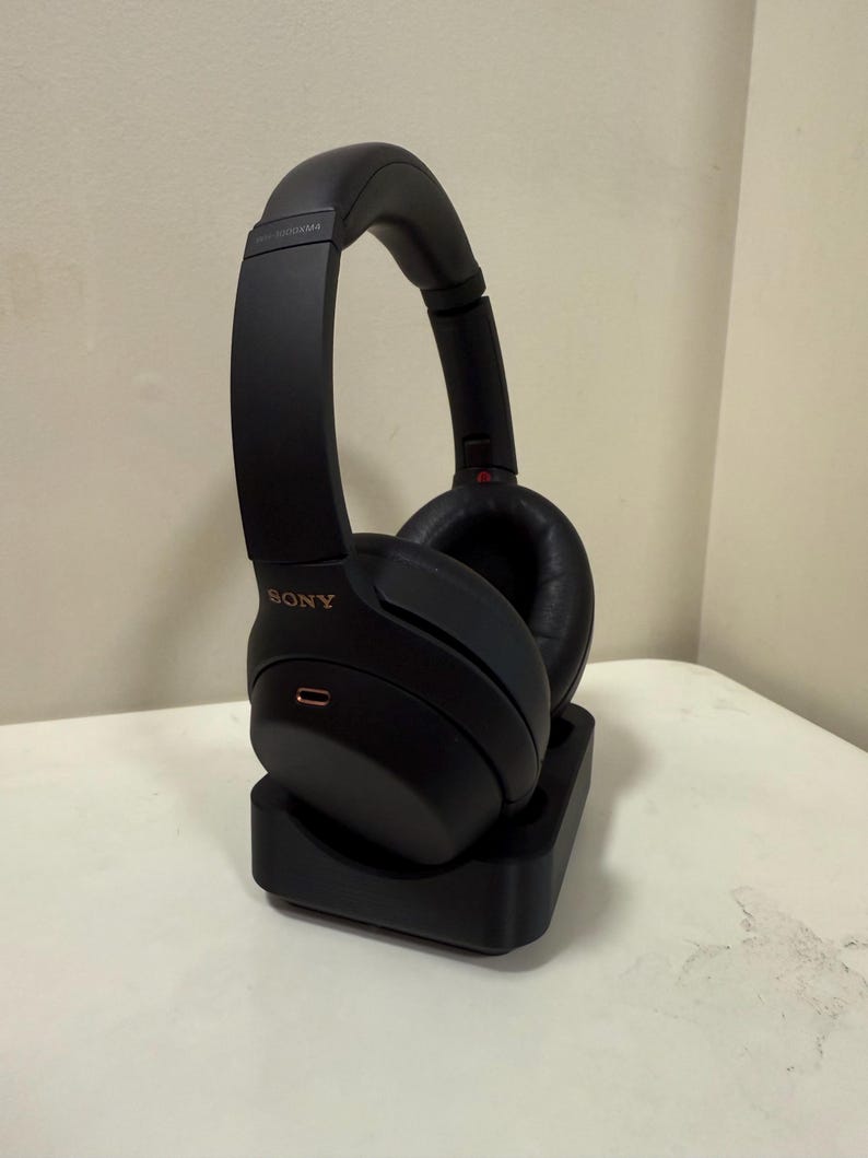 May include: Black over-ear headphones with the brand name "SONY" in gold lettering. The headphones are on a black charging stand. The earcups are round and padded. The headphones are on a white surface.