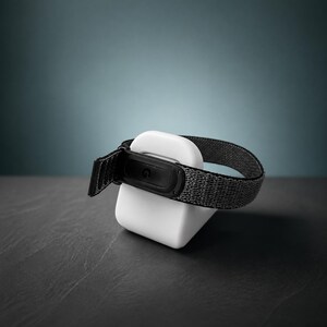 May include: A wearable fitness tracker with a dark gray woven fabric strap and a black plastic sensor module featuring a small G logo, resting on a white, rounded, square-shaped charging dock base with a smooth, matte finish and curved edges.