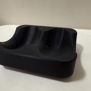 May include: A black, rectangular object with three curved indentations. The item appears to be made of a solid material and is designed to hold multiple items. The surface is smooth and the overall design is minimalist.