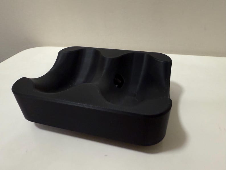 May include: A black, rectangular object with three curved indentations. The item appears to be made of a solid material and is designed to hold multiple items. The surface is smooth and the overall design is minimalist.
