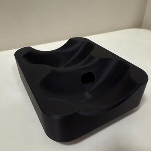 May include: A black, rectangular object with curved indentations and a central hole. The item has rounded corners and a smooth, matte finish. The design suggests it may be a holder or stand for an item.