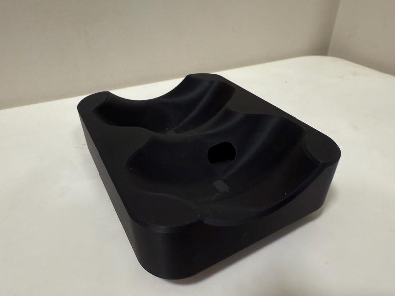 May include: A black, rectangular object with curved indentations and a central hole. The item has rounded corners and a smooth, matte finish. The design suggests it may be a holder or stand for an item.