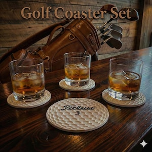Op de afbeelding: Golf Coaster Set. Drie glazen amberkleurige vloeistof met ijs staan op witte onderzetters. Een golfbalvormige onderzetter met "Titleist 3" staat op de voorgrond. Een bruine golftas en clubs staan op de achtergrond.