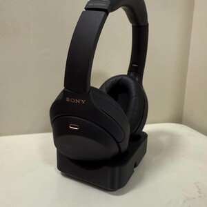 May include: Black over-ear headphones with the brand name "SONY" in gold lettering. The headphones are on a black charging stand. The earcups are made of a soft material. The headphones are designed for comfort and noise cancellation.