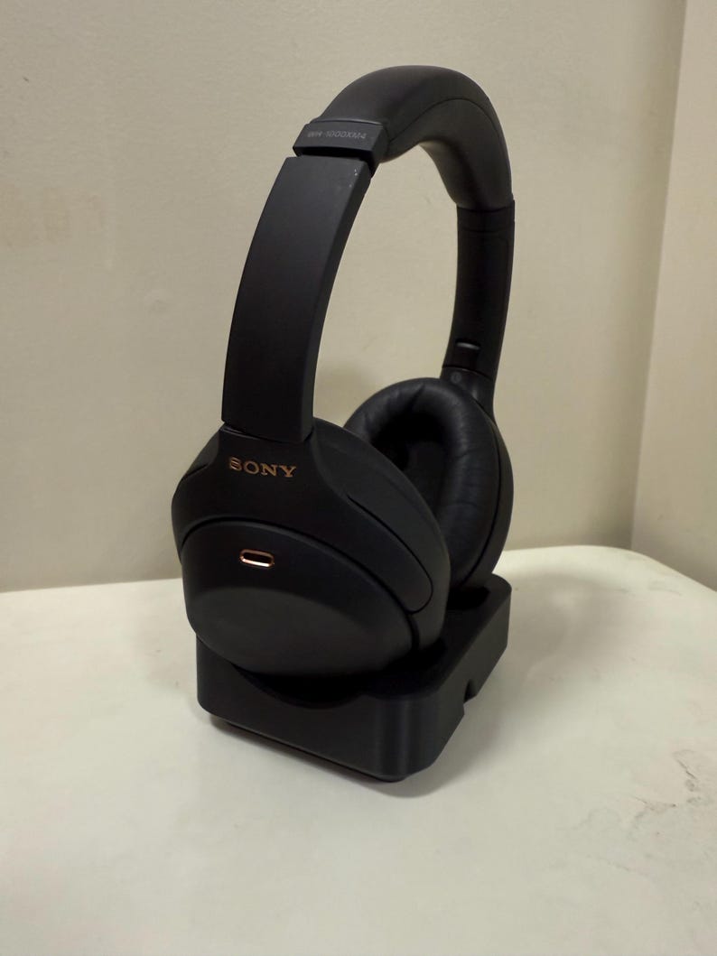 May include: Black over-ear headphones with the brand name "SONY" in gold lettering. The headphones are on a black charging stand. The earcups are made of a soft material. The headphones are designed for comfort and noise cancellation.