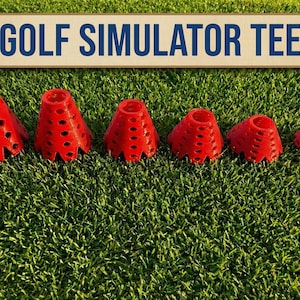 May include: A set of six red golf simulator tees of varying sizes, arranged on a bed of green artificial turf. The tees have a conical shape with multiple holes. A sign above reads "GOLF SIMULATOR TEE" in blue letters.