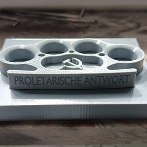 May include: A gray, 3D-printed object resembling brass knuckles, mounted on a rectangular base. The text "PROLETARISCHE ANTWORT" is embossed on the front. The design includes a hammer and sickle symbol.