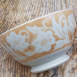 Può includere: Una piccola ciotola in ceramica bianco sporco con un motivo floreale. La ciotola ha una fascia marrone chiaro attorno al bordo con motivi floreali e di foglie bianche. La ciotola poggia su una piccola base arrotondata. Lo sfondo è una superficie di legno.